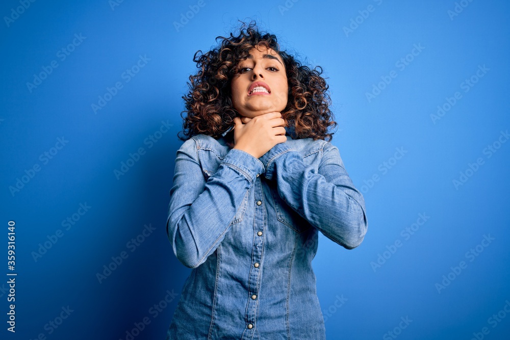 Young beautiful curly arab woman wearing casual denim shirt standing over blue background shouting and suffocate because painful strangle. Health problem. Asphyxiate and suicide concept.