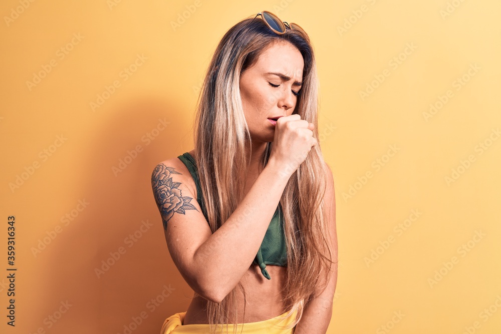 Young beautiful blonde woman wearing bikini and sunglasses standing over yellow background feeling unwell and coughing as symptom for cold or bronchitis. Health care concept.
