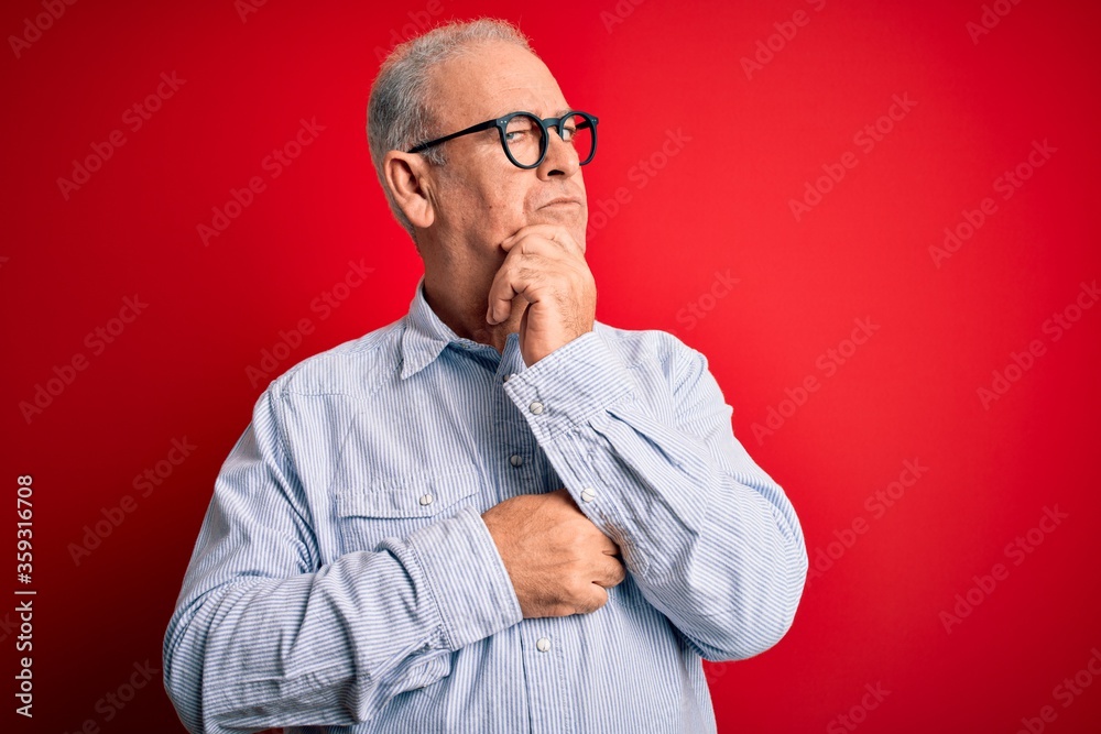 Middle age handsome hoary man wearing casual striped shirt and glasses over red background with hand on chin thinking about question, pensive expression. Smiling with thoughtful face. Doubt concept. Middle age handsome hoary man wearing casual striped shirt and glasses over red background with hand on chin thinking about question, pensive expression. Smiling with thoughtful face. Doubt concept.