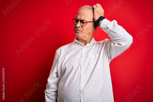 Middle age handsome hoary man wearing casual shirt and glasses over red background confuse and wondering about question. Uncertain with doubt, thinking with hand on head. Pensive concept.