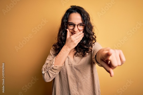 Beautiful woman with curly hair wearing striped shirt and glasses over yellow background laughing at you, pointing finger to the camera with hand over mouth, shame expression