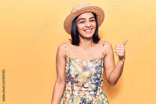Young beautiful latin woman wearing summer hat smiling with happy face looking and pointing to the side with thumb up.