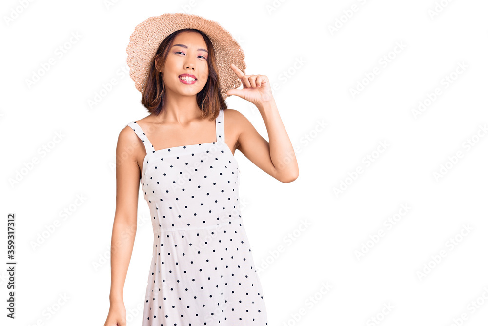 Young beautiful chinese girl wearing summer hat smiling and confident gesturing with hand doing small size sign with fingers looking and the camera. measure concept. Young beautiful chinese girl wearing summer hat smiling and confident gesturing with hand doing small size sign with fingers looking and the camera. measure concept.
