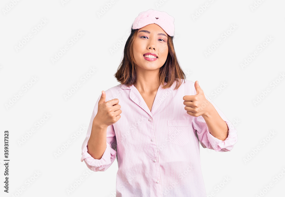 Young beautiful chinese girl wearing sleep mask and pajama success sign doing positive gesture with hand, thumbs up smiling and happy. cheerful expression and winner gesture. Young beautiful chinese girl wearing sleep mask and pajama success sign doing positive gesture with hand, thumbs up smiling and happy. cheerful expression and winner gesture.