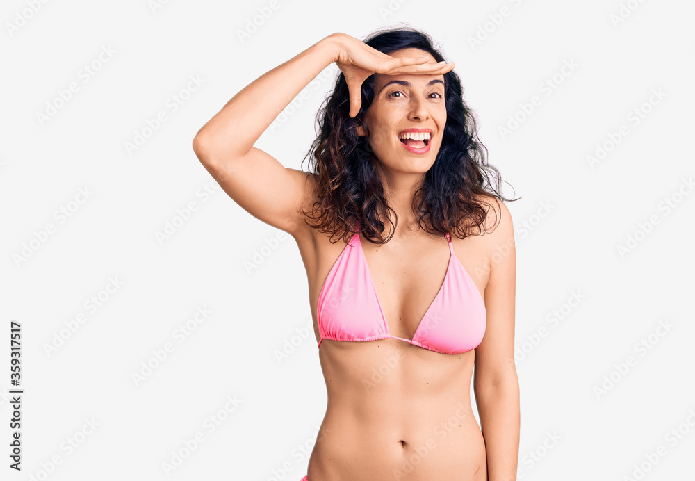 Fototapeta premium Young beautiful hispanic woman wearing bikini very happy and smiling looking far away with hand over head. searching concept.