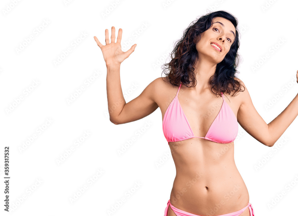 Young beautiful hispanic woman wearing bikini clueless and confused expression with arms and hands raised. doubt concept. Young beautiful hispanic woman wearing bikini clueless and confused expression with arms and hands raised. doubt concept.