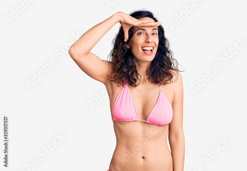 Young beautiful hispanic woman wearing bikini very happy and smiling looking far away with hand over head. searching concept. © Krakenimages.com