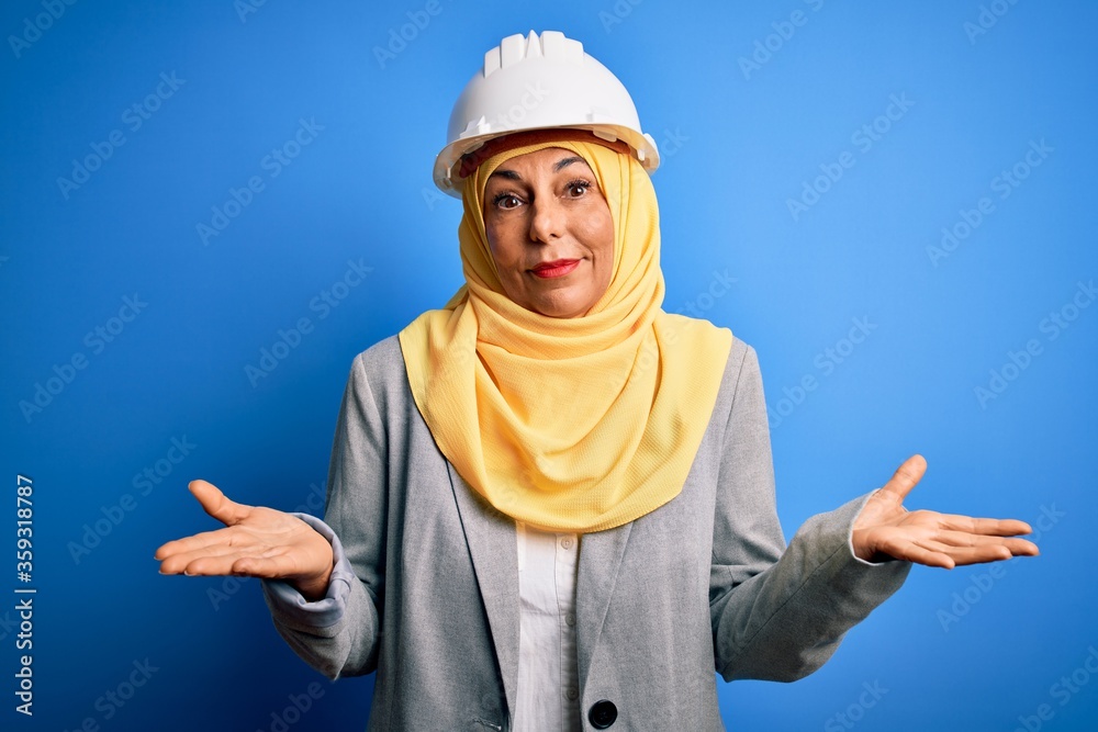Middle age brunette architect woman wearing muslim traditional hijab and security helmet clueless and confused expression with arms and hands raised. Doubt concept. Middle age brunette architect woman wearing muslim traditional hijab and security helmet clueless and confused expression with arms and hands raised. Doubt concept.