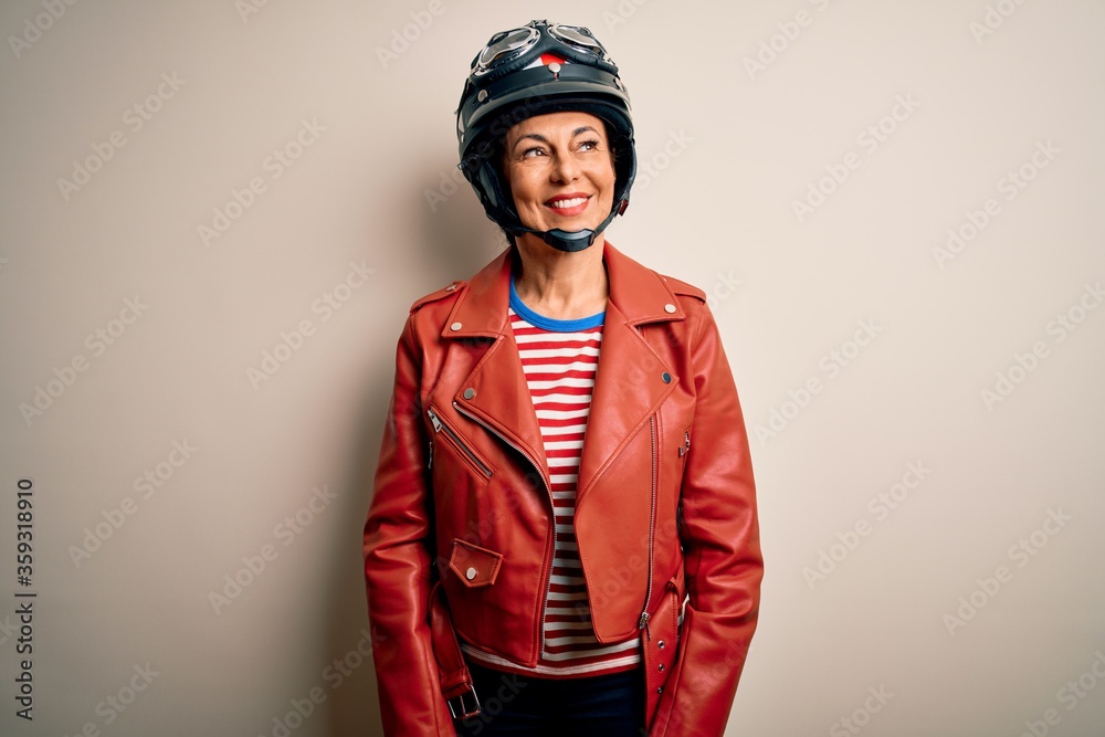 Middle age motorcyclist woman wearing motorcycle helmet and jacket over white background looking away to side with smile on face, natural expression. Laughing confident. Middle age motorcyclist woman wearing motorcycle helmet and jacket over white background looking away to side with smile on face, natural expression. Laughing confident.