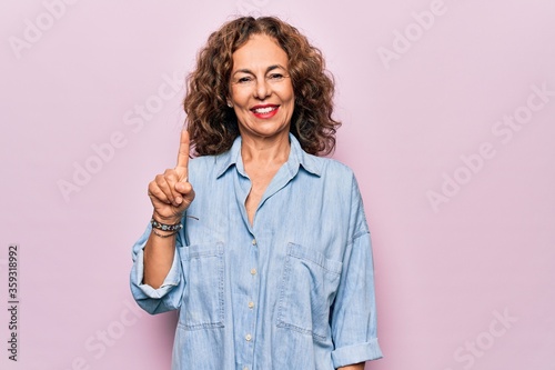 Middle age beautiful woman wearing casual denim shirt standing over pink background pointing finger up with successful idea. Exited and happy. Number one.