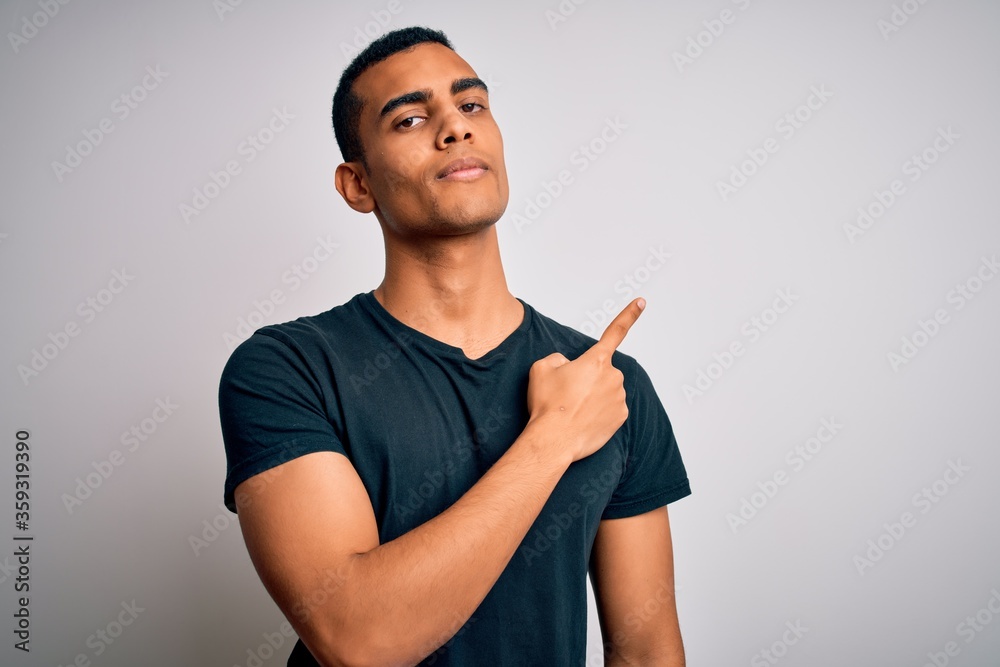Young handsome african american man wearing casual t-shirt standing over white background Pointing with hand finger to the side showing advertisement, serious and calm face Young handsome african american man wearing casual t-shirt standing over white background Pointing with hand finger to the side showing advertisement, serious and calm face