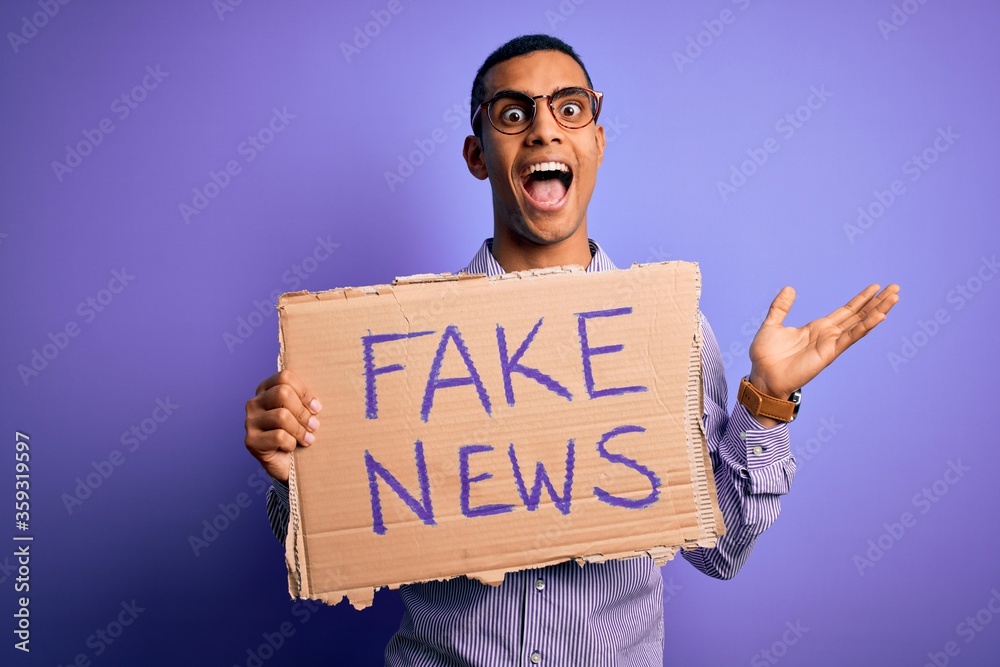 Young handsome african american man protesting holding banner with fake news message very happy and excited, winner expression celebrating victory screaming with big smile and raised hands Young handsome african american man protesting holding banner with fake news message very happy and excited, winner expression celebrating victory screaming with big smile and raised hands