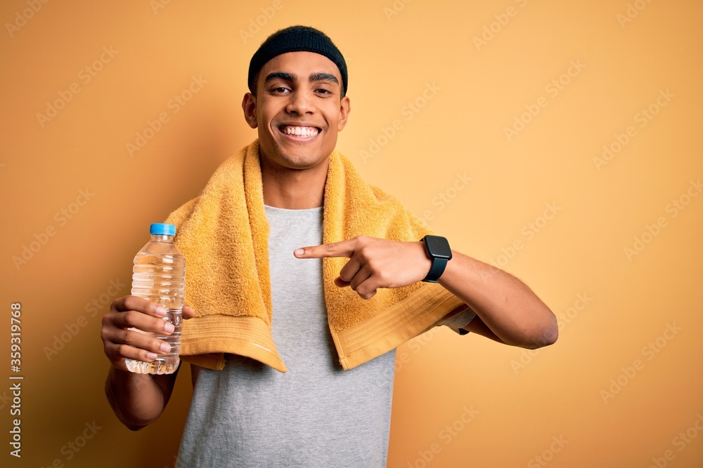 Young handsome african american sportsman wearing towel drinking bottle of water very happy pointing with hand and finger Young handsome african american sportsman wearing towel drinking bottle of water very happy pointing with hand and finger