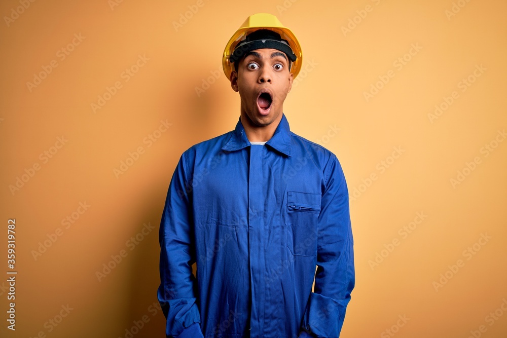 Young handsome african american worker man wearing blue uniform and security helmet afraid and shocked with surprise and amazed expression, fear and excited face. Young handsome african american worker man wearing blue uniform and security helmet afraid and shocked with surprise and amazed expression, fear and excited face.