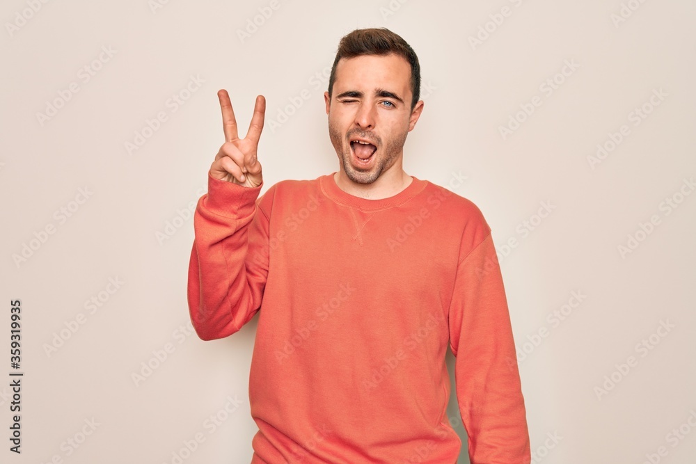 Fototapeta premium Young handsome man with blue eyes wearing casual sweater standing over white background smiling with happy face winking at the camera doing victory sign. Number two.