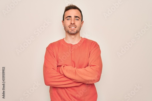 Young handsome man with blue eyes wearing casual sweater standing over white background happy face smiling with crossed arms looking at the camera. Positive person.