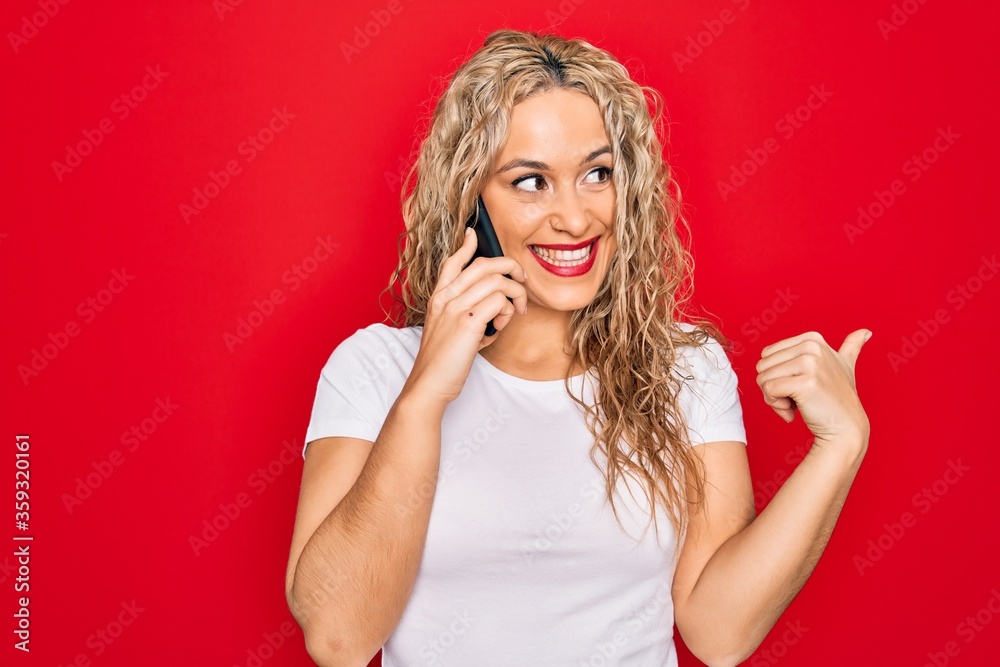 Beautiful blonde woman having conversation talking on the smartphone over red background pointing thumb up to the side smiling happy with open mouth Beautiful blonde woman having conversation talking on the smartphone over red background pointing thumb up to the side smiling happy with open mouth