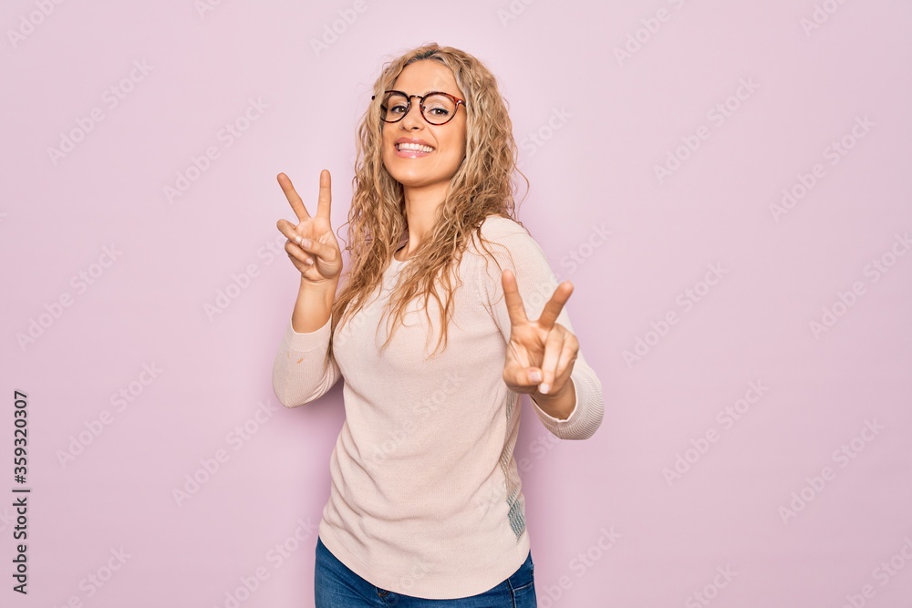 Φωτογραφία Young beautiful blonde woman wearing casual sweater and glasses over pink background smiling looking to the camera showing fingers doing victory sign