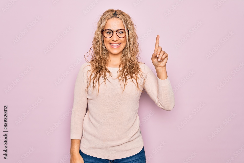 Young beautiful blonde woman wearing casual sweater and glasses over pink background showing and pointing up with finger number one while smiling confident and happy. Young beautiful blonde woman wearing casual sweater and glasses over pink background showing and pointing up with finger number one while smiling confident and happy.