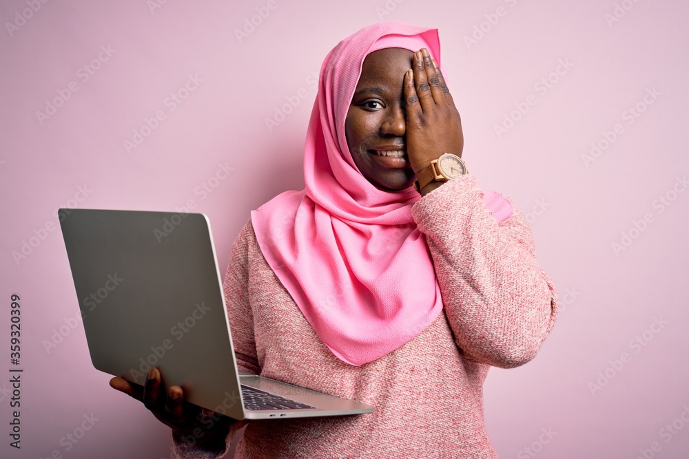 African american plus size woman wearing muslim hijab using laptop over pink background covering one eye with hand, confident smile on face and surprise emotion. African american plus size woman wearing muslim hijab using laptop over pink background covering one eye with hand, confident smile on face and surprise emotion.