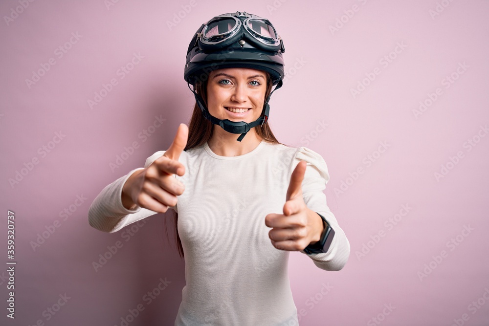 Young beautiful motorcyclist woman with blue eyes wearing moto helmet over pink background pointing fingers to camera with happy and funny face. Good energy and vibes. Young beautiful motorcyclist woman with blue eyes wearing moto helmet over pink background pointing fingers to camera with happy and funny face. Good energy and vibes.