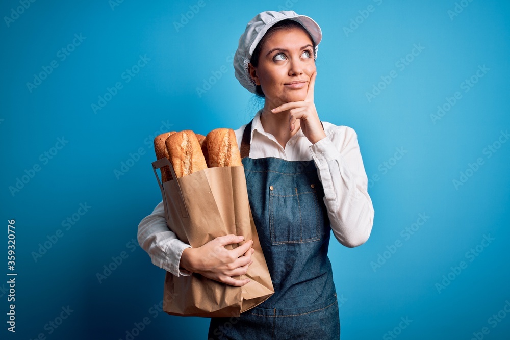 Young beautiful baker woman with blue eyes holding paper bag with  bread serious face thinking about question, very confused idea