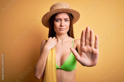 Young beautiful woman with blue eyes on vacation wearing bikini and hat holding towel with open hand doing stop sign with serious and confident expression, defense gesture