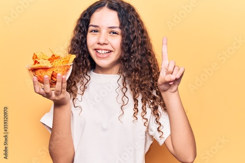 Beautiful kid girl with curly hair holding nachos potato chips smiling with an idea or question pointing finger with happy face, number one