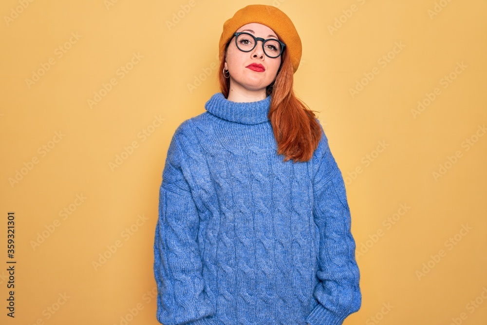 Obraz premium Young beautiful redhead woman wearing french beret and glasses over yellow background Relaxed with serious expression on face. Simple and natural looking at the camera.