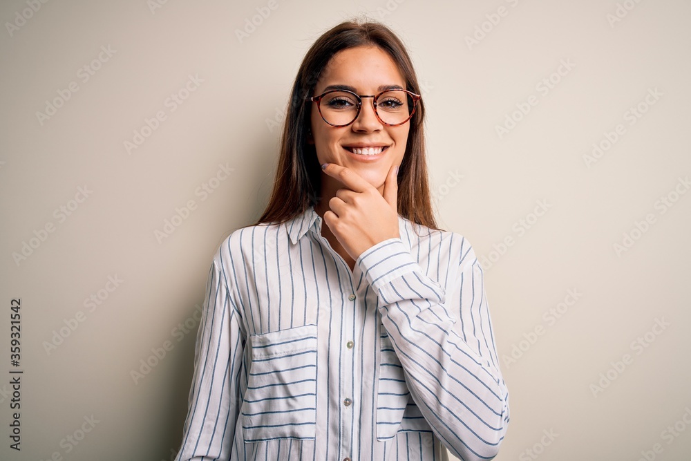 Young beautiful brunette woman wearing casual shirt and glasses over white background looking confident at the camera smiling with crossed arms and hand raised on chin. Thinking positive.