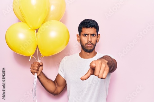 Young latin man holding balloons pointing with finger to the camera and to you, confident gesture looking serious © Krakenimages.com
