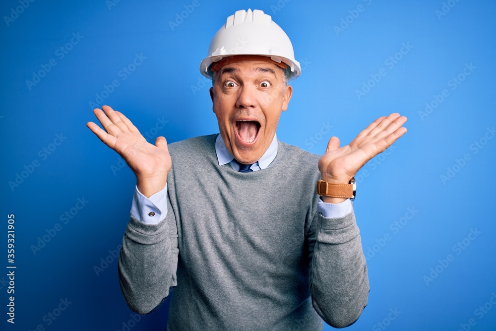 Middle age handsome grey-haired engineer man wearing safety helmet over blue background celebrating crazy and amazed for success with arms raised and open eyes screaming excited. Winner concept Middle age handsome grey-haired engineer man wearing safety helmet over blue background celebrating crazy and amazed for success with arms raised and open eyes screaming excited. Winner concept