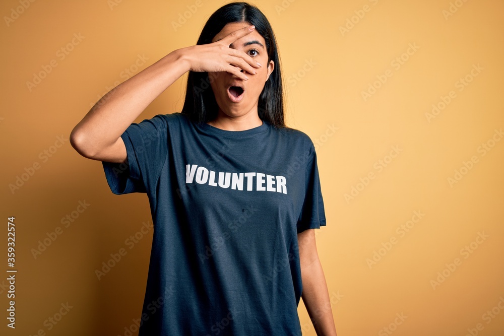 Young beautiful hispanic woman wearing volunteer t-shirt as social charity moral peeking in shock covering face and eyes with hand, looking through fingers with embarrassed expression. Young beautiful hispanic woman wearing volunteer t-shirt as social charity moral peeking in shock covering face and eyes with hand, looking through fingers with embarrassed expression.