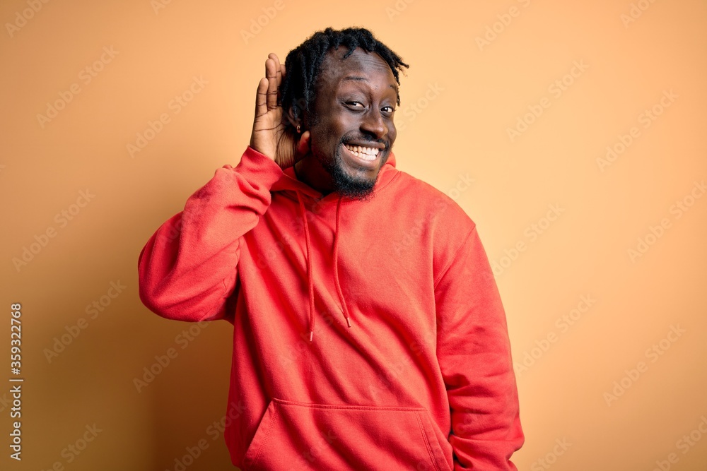 Young african american sporty man wearing sweatshirt with hoodie over yellow background smiling with hand over ear listening an hearing to rumor or gossip. Deafness concept.