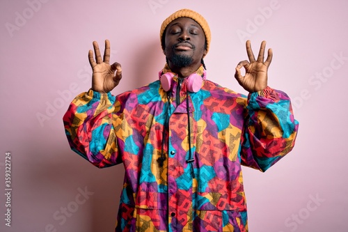 Young handsome african american man wearing colorful coat and cap over pink background relax and smiling with eyes closed doing meditation gesture with fingers. Yoga concept.