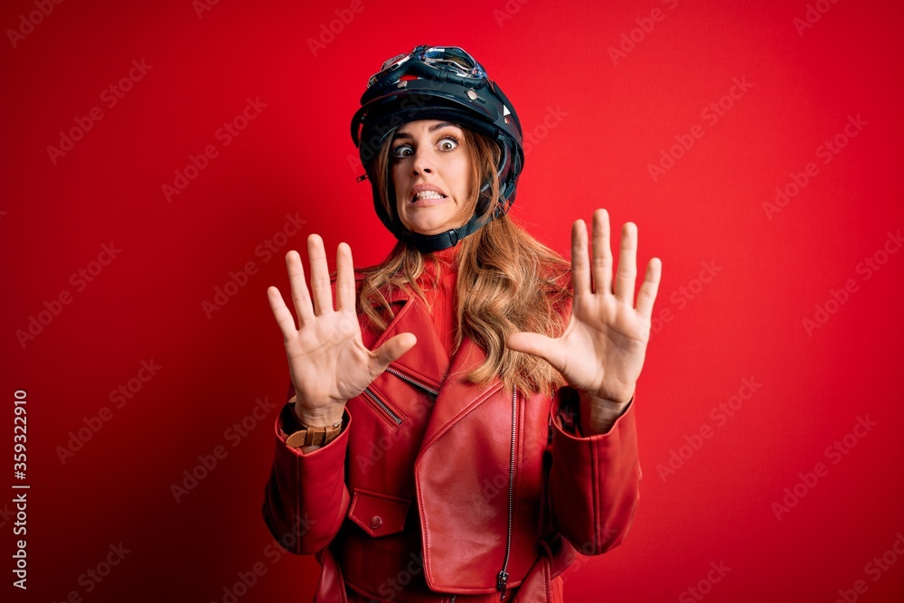 Young beautiful brunette motrocyclist woman wearing moto helmet over red background afraid and terrified with fear expression stop gesture with hands, shouting in shock. Panic concept. Young beautiful brunette motrocyclist woman wearing moto helmet over red background afraid and terrified with fear expression stop gesture with hands, shouting in shock. Panic concept.