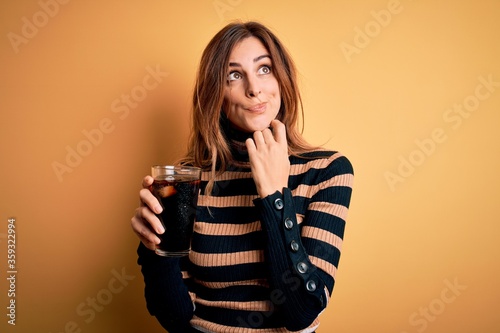 Young beautiful brunette woman drinking glass with cola refreshment using straw serious face thinking about question, very confused idea