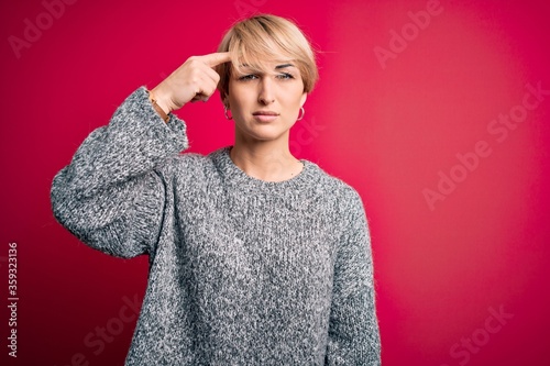 Young blonde woman with modern short hair wearing casual sweater over pink background pointing unhappy to pimple on forehead, ugly infection of blackhead. Acne and skin problem © Krakenimages.com