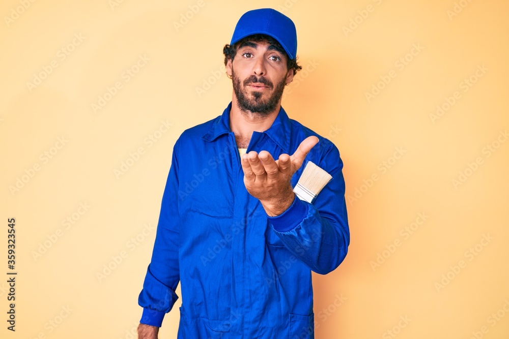 Handsome young man with curly hair and bear wearing builder jumpsuit uniform looking at the camera blowing a kiss with hand on air being lovely and sexy. love expression.