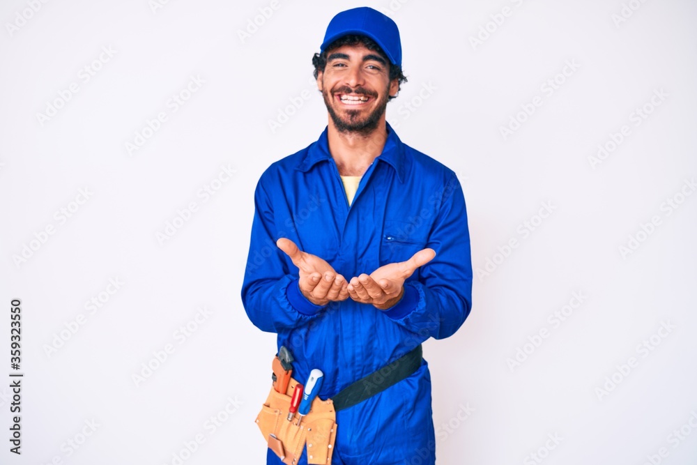 Handsome young man with curly hair and bear weaing handyman uniform smiling with hands palms together receiving or giving gesture. hold and protection Handsome young man with curly hair and bear weaing handyman uniform smiling with hands palms together receiving or giving gesture. hold and protection