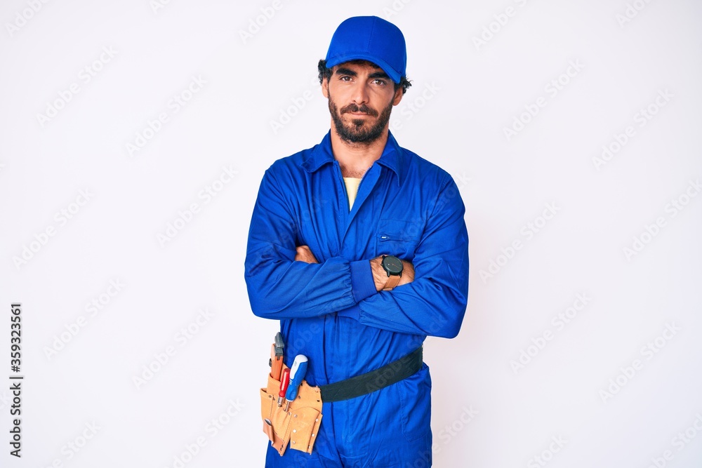 Handsome young man with curly hair and bear weaing handyman uniform skeptic and nervous, disapproving expression on face with crossed arms. negative person.