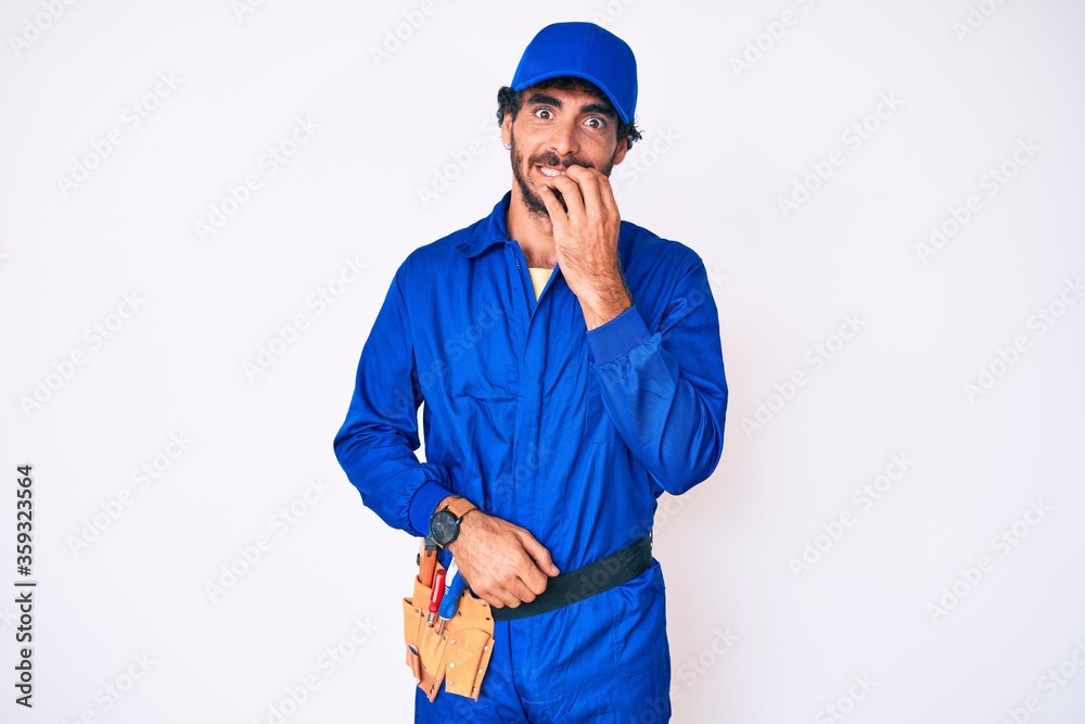 Handsome young man with curly hair and bear weaing handyman uniform looking stressed and nervous with hands on mouth biting nails. anxiety problem.