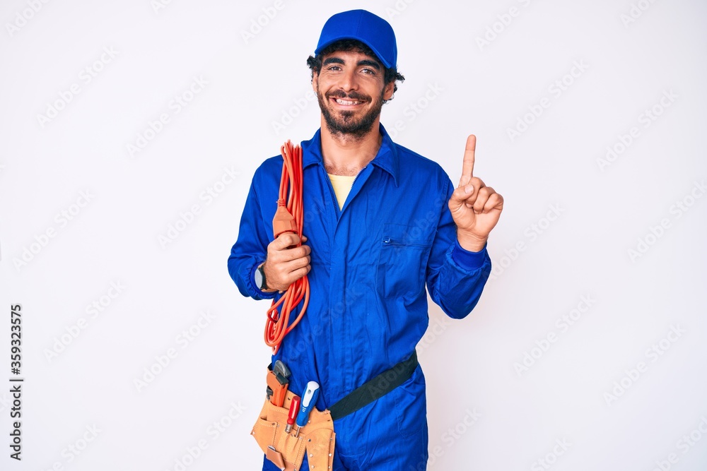 Handsome young man with curly hair and bear wearing builder jumpsuit uniform and electric cables smiling with an idea or question pointing finger with happy face, number one