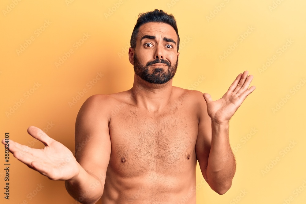 Young hispanic man standing shirtless clueless and confused expression with arms and hands raised. doubt concept.