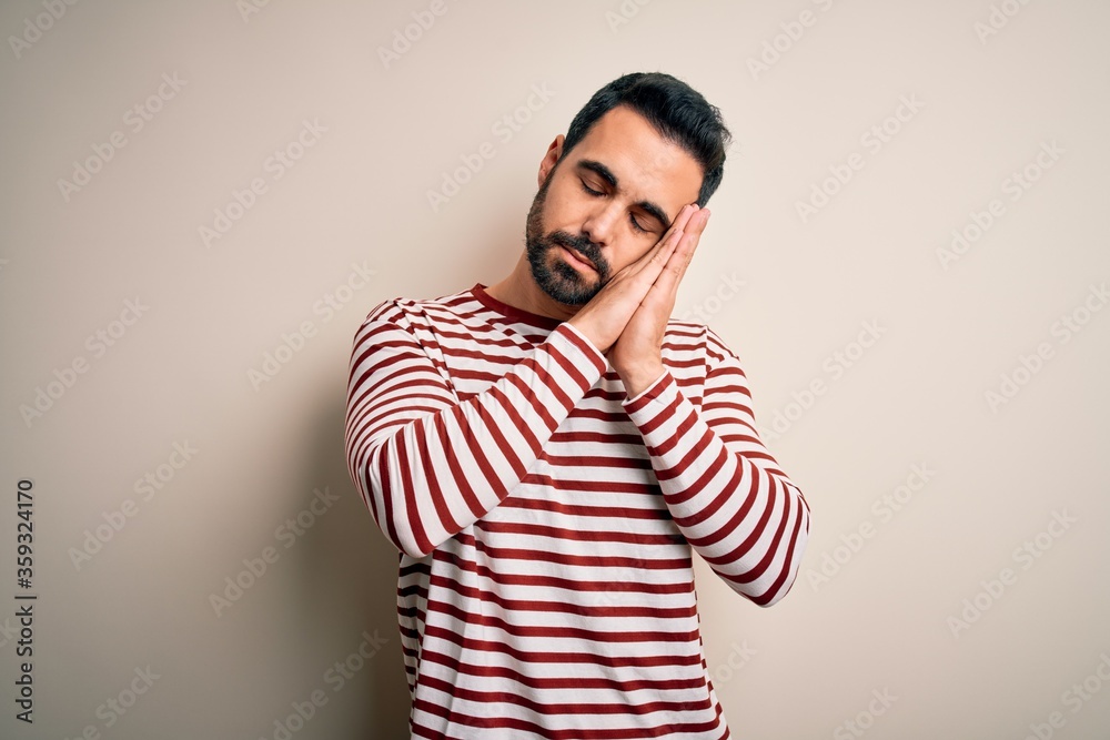 Fototapeta premium Young handsome man with beard wearing casual striped t-shirt standing over white background sleeping tired dreaming and posing with hands together while smiling with closed eyes.