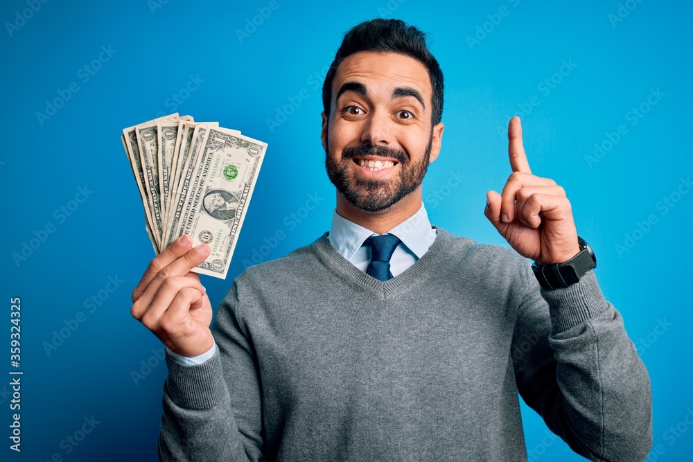 Young handsome man with beard holding bunch of dollars banknotes over blue background surprised with an idea or question pointing finger with happy face, number one Young handsome man with beard holding bunch of dollars banknotes over blue background surprised with an idea or question pointing finger with happy face, number one