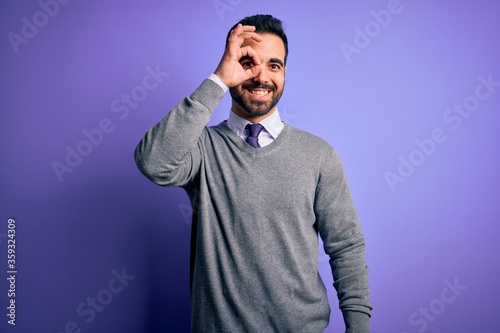 Handsome businessman with beard wearing casual tie standing over purple background doing ok gesture with hand smiling, eye looking through fingers with happy face.