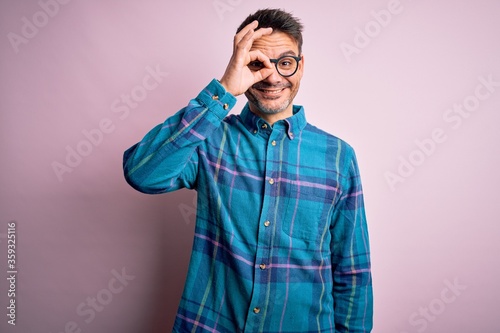 Young handsome man wearing casual shirt and glasses standing over isolated pink background doing ok gesture with hand smiling, eye looking through fingers with happy face. © Krakenimages.com