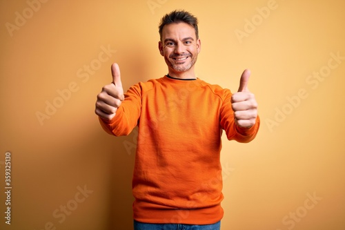 Young handsome man wearing orange casual sweater standing over isolated yellow background approving doing positive gesture with hand, thumbs up smiling and happy for success. Winner gesture.