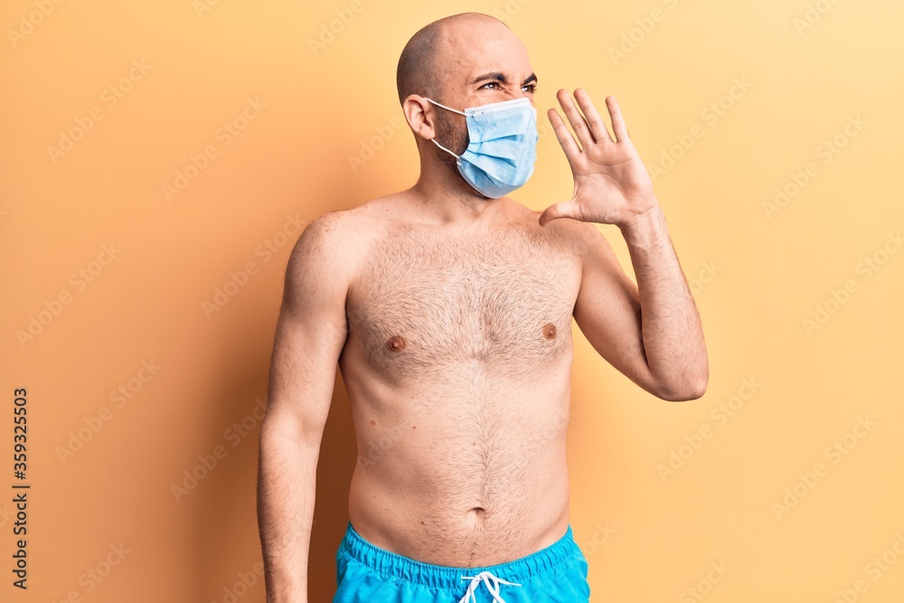 Fototapeta premium Young handsome bald man shirtless wearing swimwear and medical mask shouting and screaming loud to side with hand on mouth. communication concept.
